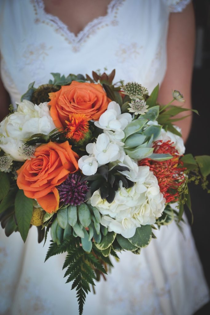 Beautiful large fall floral bouquet with orange roses and greenery being held at waist height by a vintage inspired bride.
