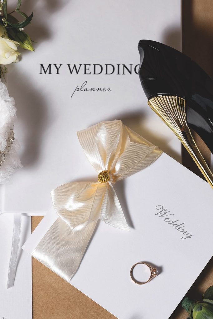 A wedding planner sits on a table with a ring and a pair of shoes. The planner is white and gold, and the ring is gold and silver. Concept of elegance and sophistication